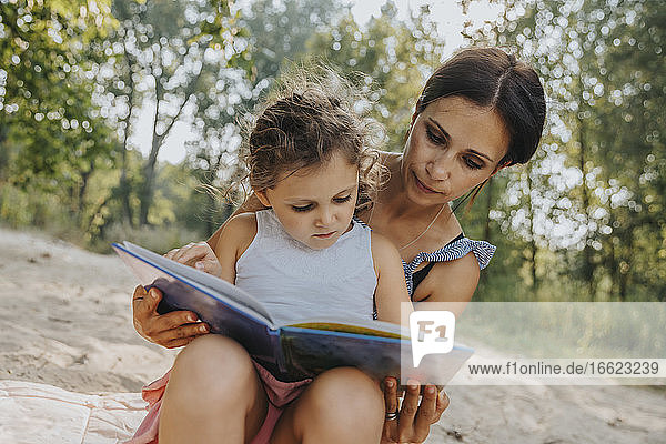 Mother and daughter reading book while sitting at beach