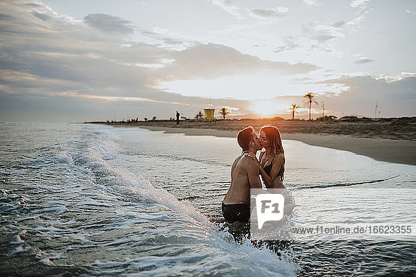 Paar in Badekleidung küsst sich bei Sonnenuntergang am Strand sitzend