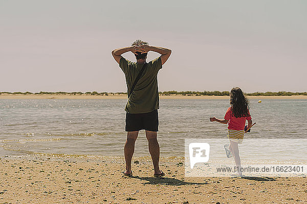 Father with daughter standing at beach against clear sky during sunny day