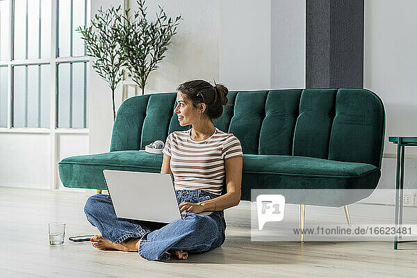 Nachdenkliche Architektin  die im Schneidersitz auf dem Boden sitzt und einen Laptop auf dem Sofa im Büro benutzt