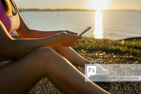 Nahaufnahme einer jungen Frau  die ihr Smartphone benutzt  während sie bei Sonnenuntergang auf einer Straße am Meer sitzt