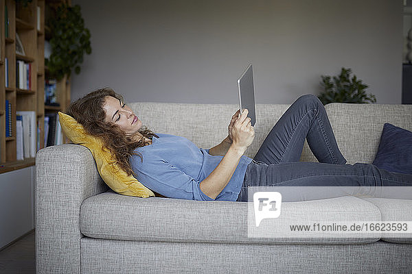 Frau benutzt digitales Tablet  während sie zu Hause auf dem Sofa liegt