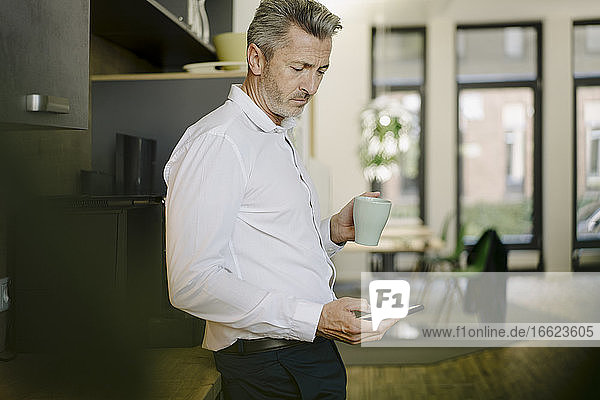 Geschäftsmann hält Tasse  während er ein Mobiltelefon im Büro benutzt