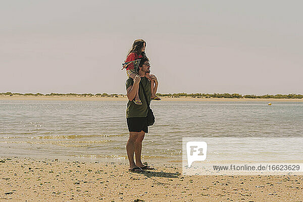 Father carrying daughter on shoulders while standing at beach against clear sky during sunny day