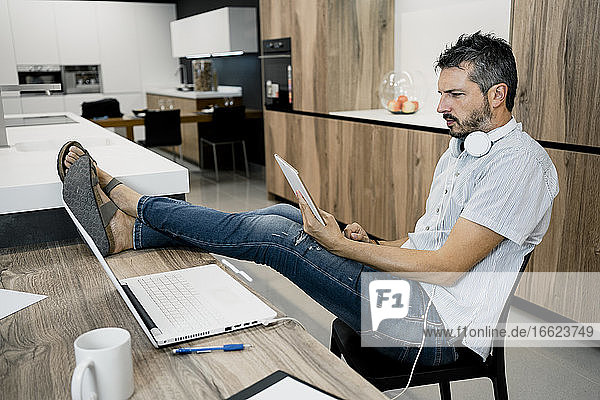 Mature male freelancer holding digital tablet while sitting with feet up on desk at office