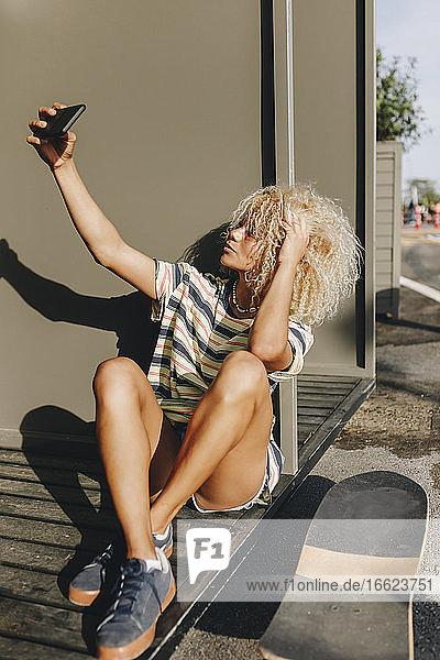 Blonde Frau  die ein Selfie mit ihrem Smartphone macht  während sie an einer Metallwand sitzt