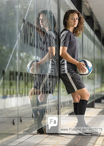 Eine Fußballspielerin hält einen Ball und steht vor einer Glaswand