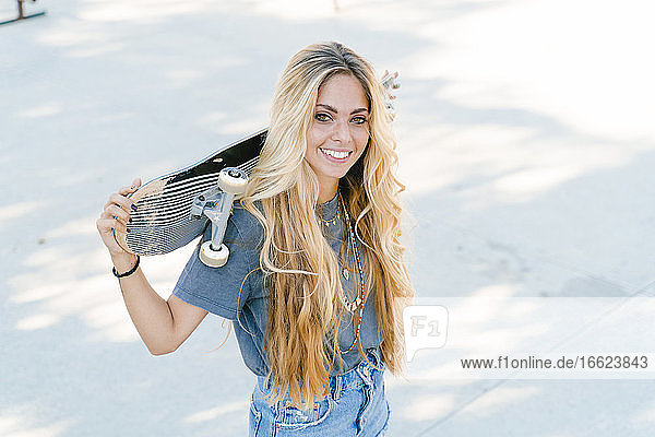Lächelnde blonde Frau  die ein Skateboard hält  während sie im Park steht