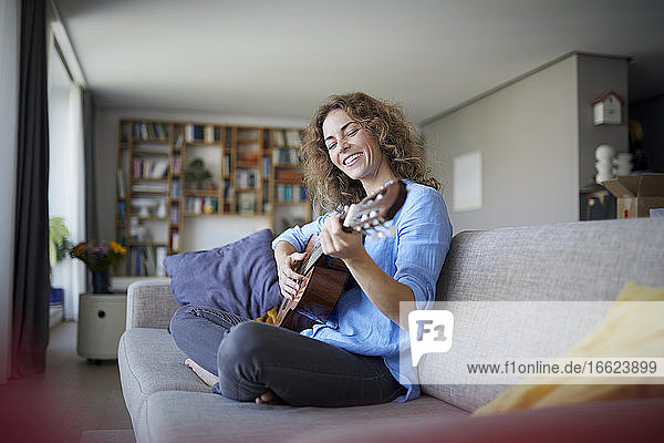 Lächelnde Frau spielt Gitarre  während sie zu Hause auf dem Sofa sitzt