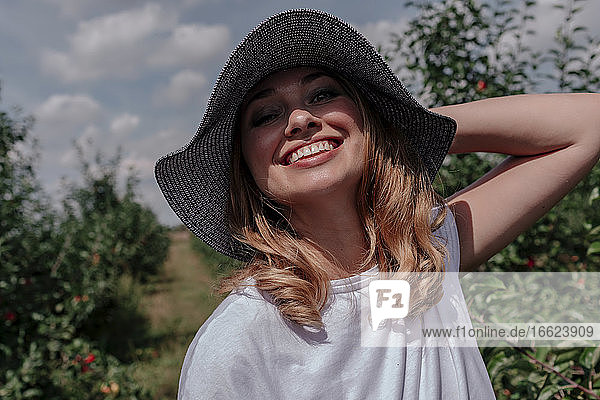 Nahaufnahme einer lächelnden erwachsenen Frau mit Hut in einem Obstgarten