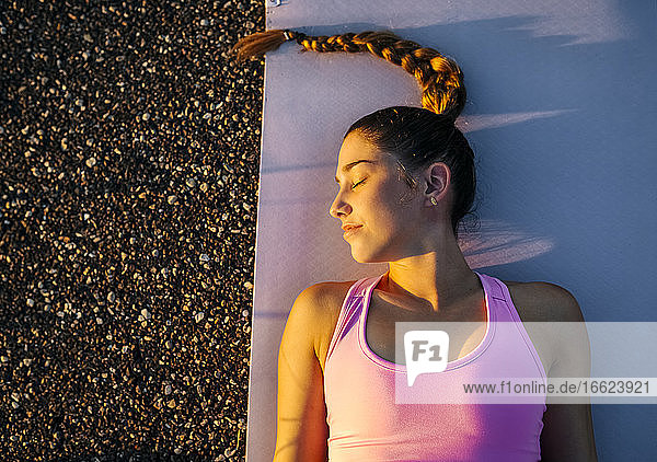 Junge Frau mit geschlossenen Augen meditiert im Liegen auf einer Matte bei Sonnenuntergang