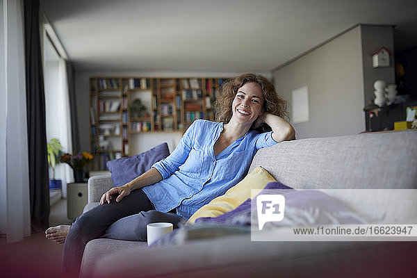 Lächelnde Frau mit dem Kopf in den Händen sitzt zu Hause auf dem Sofa