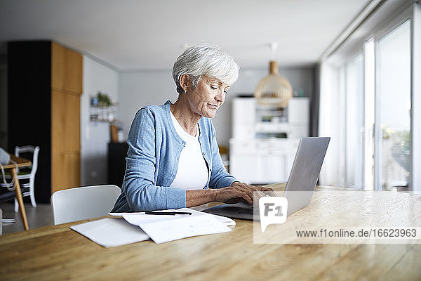 Ältere Frau arbeitet am Laptop  während sie zu Hause auf einem Stuhl sitzt