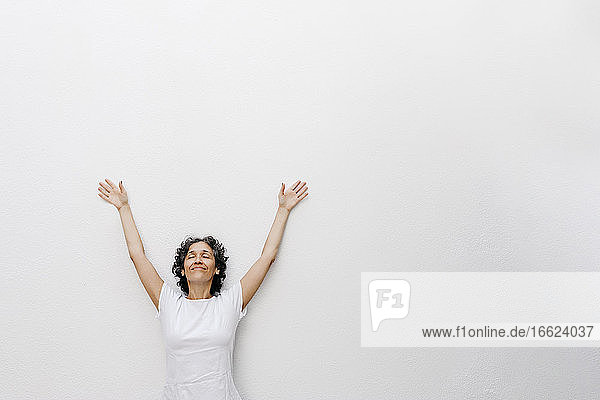 Lächelnde reife Frau steht mit geschlossenen Augen und erhobenen Armen vor einer weißen Wand