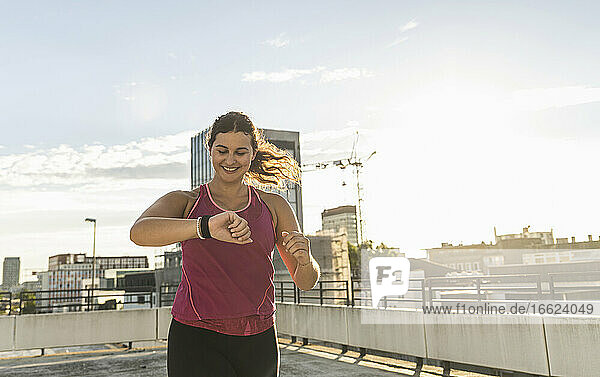 Lächelnde junge Frau prüft die Zeit beim Laufen auf der Terrasse gegen den Himmel