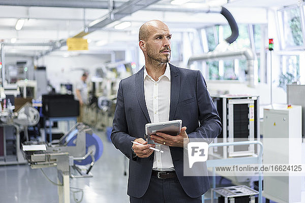 Nachdenklicher Geschäftsmann hält ein digitales Tablet in der Hand und blickt auf eine beleuchtete Fabrik
