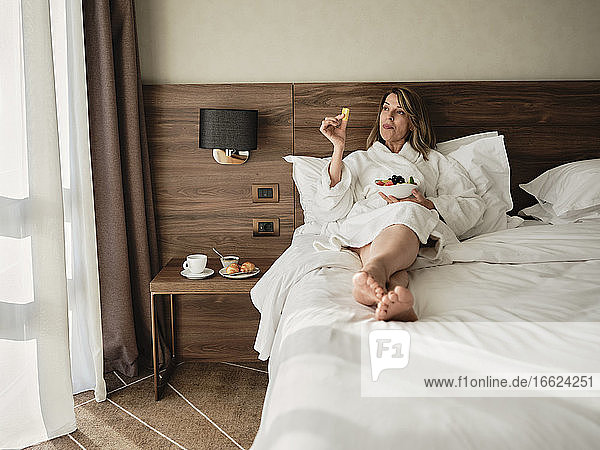 Senior woman eating fresh fruits in breakfast while relaxing on bed and looking away at hotel room