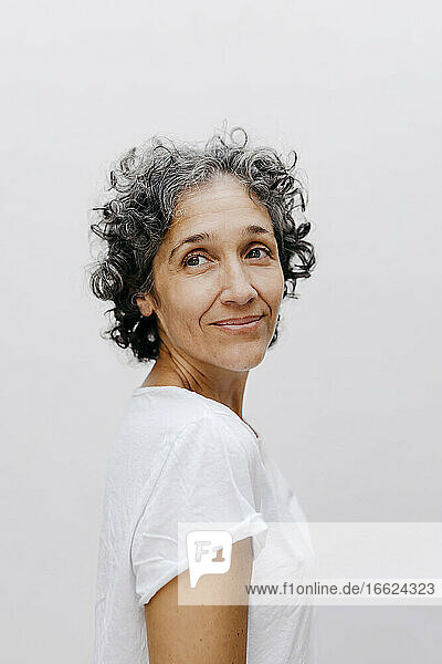 Mature woman with short curly hair looking away while standing against white wall