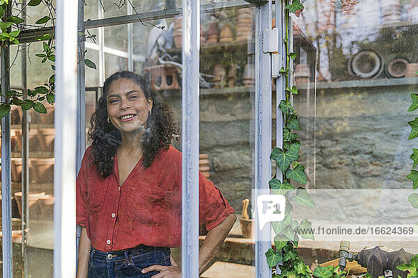 Reife Frau mit Hand auf der Hüfte  die durch ein Fenster schaut  während sie in einem Gartenhaus steht
