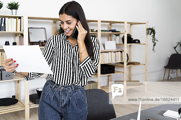 Lächelnde junge Architektin  die mit ihrem Smartphone spricht  während sie einen Plan im Büro betrachtet