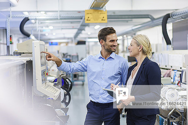 Lächelnder männlicher Techniker  der eine Geschäftsfrau ansieht  während er in einer beleuchteten Fabrik vor Maschinen steht