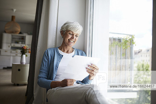 Ältere Frau liest fröhlich einen Brief  während sie zu Hause am Fenster sitzt