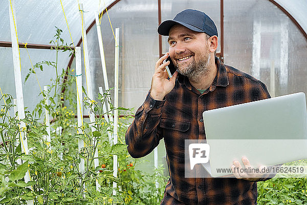 Man holding laptop while talking on mobile phone at greenhouse