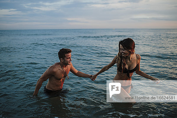 Paar hält sich an der Hand  während es bei Sonnenuntergang am Strand im Wasser spazieren geht