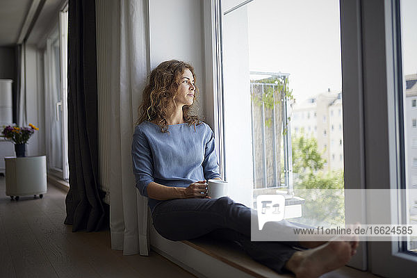 Frau hält Kaffeetasse  während sie auf der Fensterbank zu Hause sitzt