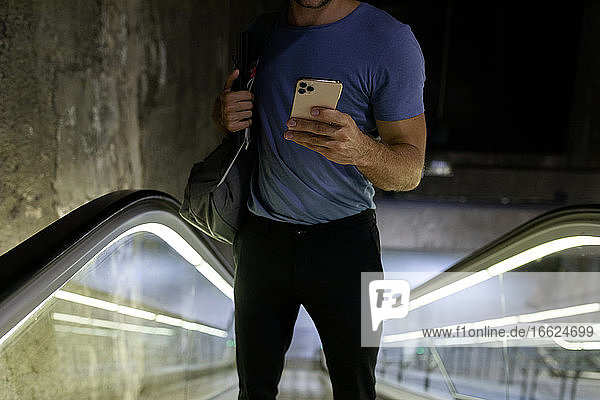 Junger Mann benutzt sein Smartphone  während er mit seinem Rucksack auf einer Rolltreppe in der U-Bahn steht
