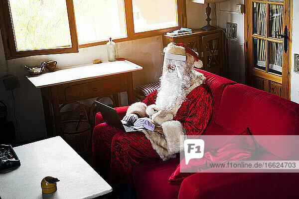 Mann im Weihnachtsmannkostüm benutzt Laptop  während er zu Hause auf dem Sofa sitzt