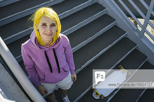 Dyed hair hipster female with skateboard standing on staircase during sunny day