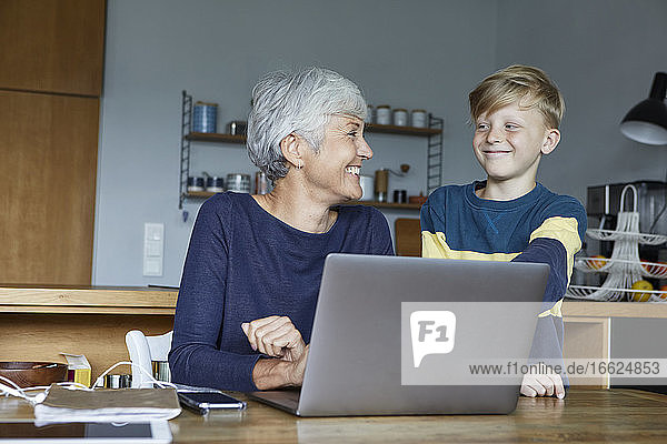 Lächelnder Enkel steht neben der Großmutter  die zu Hause am Laptop arbeitet