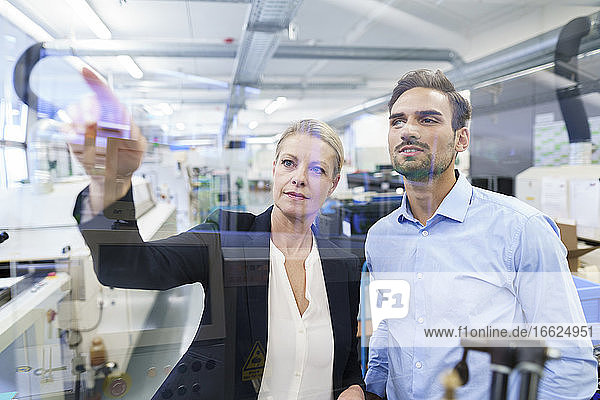 Selbstbewusste Geschäftsfrau  die mit einem jungen Ingenieur plant  während sie auf eine Glasoberfläche in einer Fabrik zeigt