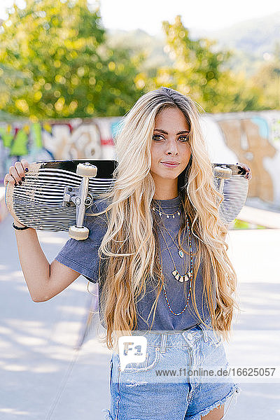 Selbstbewusste blonde Frau mit Skateboard im Park stehend