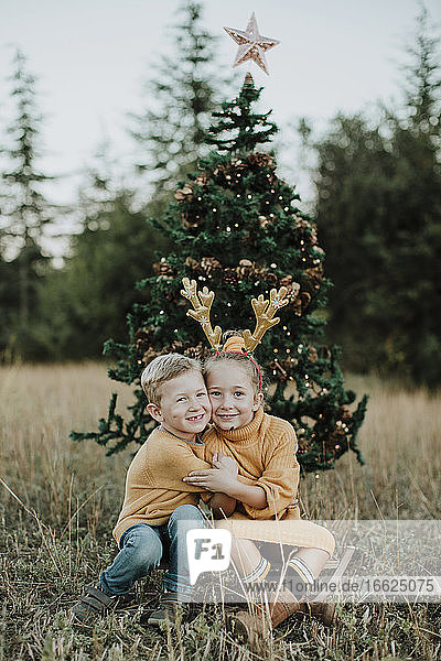 Lächelnde Geschwister  die sich umarmen  während sie am Weihnachtsbaum auf einer Wiese sitzen
