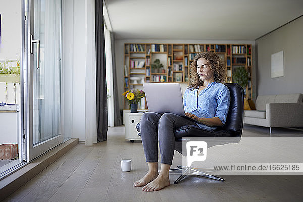 Mittlere erwachsene Frau arbeitet am Laptop  während sie zu Hause auf einem Stuhl sitzt