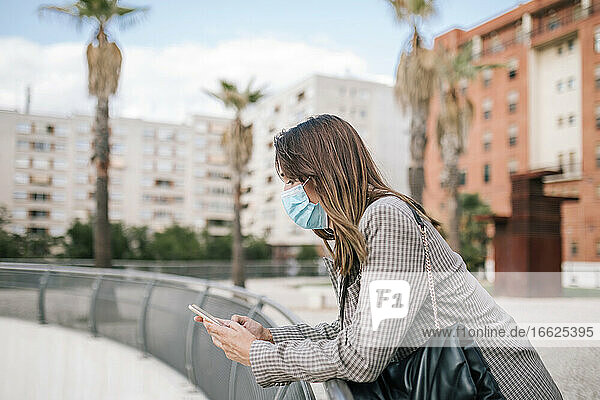 Junge Frau mit Gesichtsmaske  die ihr Smartphone benutzt und sich auf ein Geländer in der Stadt stützt