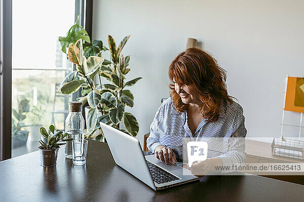 Mature woman sitting by table while working on laptop at home
