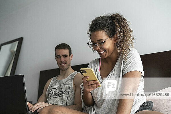 Mann arbeitet an einem Laptop und sieht eine Frau  die zu Hause ein Mobiltelefon benutzt
