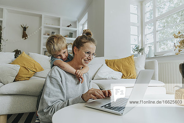 Frau arbeitet am Laptop  während ein Junge sie zu Hause von hinten umarmt