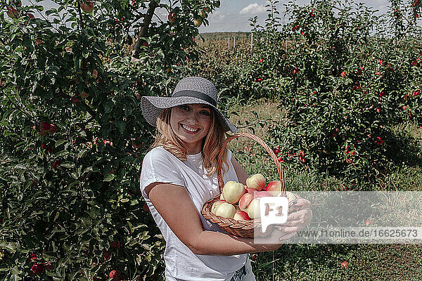 Smiling woman wearing hat holding apples in basket while standing at orchard