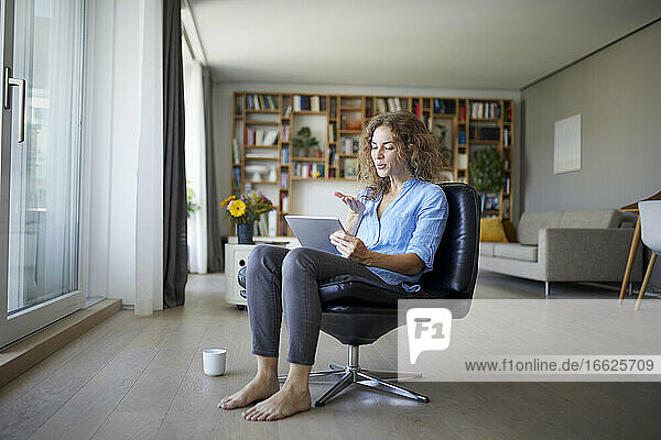 Frau bläst Kuss zum Videoanruf auf digitalem Tablet  während sie zu Hause auf einem Stuhl sitzt
