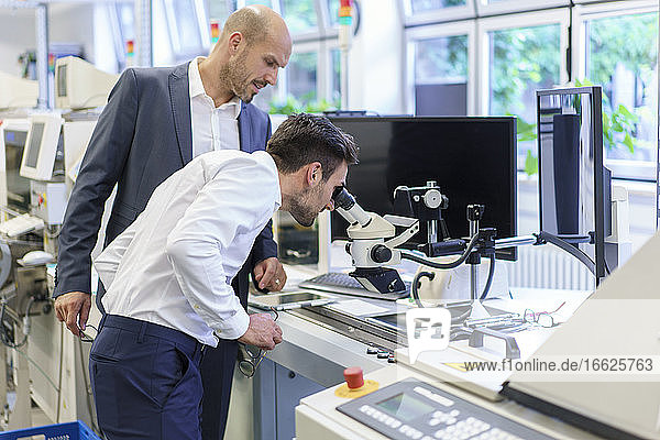 Junger Ingenieur schaut durch ein Mikroskop  während er neben einem Geschäftsmann im Labor steht