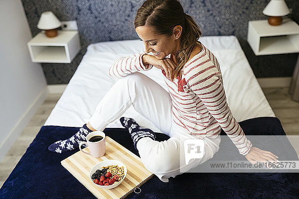 Lächelnde Frau  die sich Essen und Kaffee ansieht  während sie zu Hause auf dem Bett sitzt