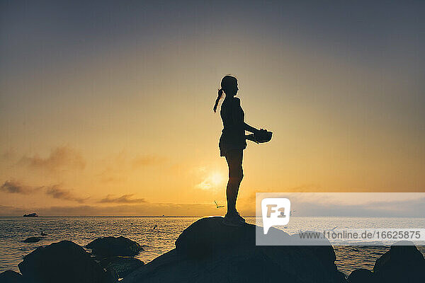 Silhouette einer Frau  die eine Schüssel hält  während sie bei Sonnenuntergang auf einem Felsen am Strand steht
