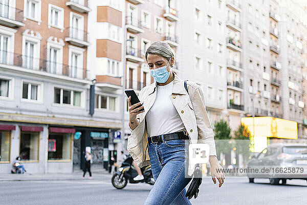 Junge Frau mit Maske  die ein Mobiltelefon benutzt  während sie auf einer Straße in der Stadt spazieren geht
