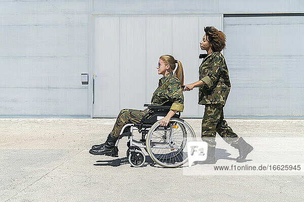 Junger weiblicher Soldat schiebt einen reifen Kollegen im Rollstuhl auf einem Armeestützpunkt an einem sonnigen Tag