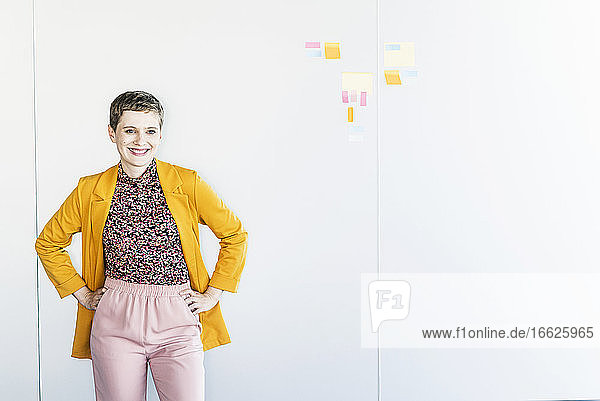 Lächelnde Unternehmerin mit Händen in den Taschen  die an einer weißen Wand im Büro steht