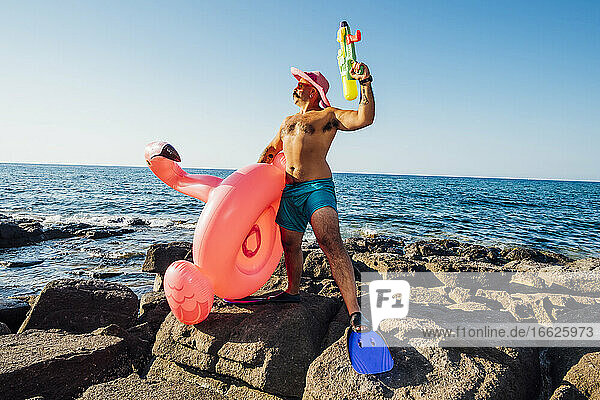 Stehender Mann  der einen Flamingo-Ring und eine Wasserpistole gegen das Meer hält